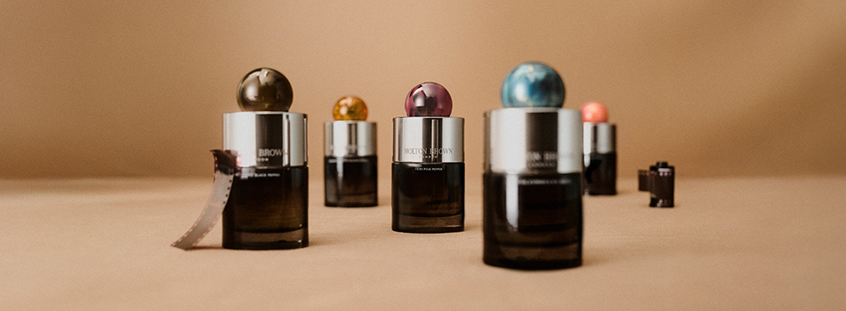 Fünf edle Parfumflakons von Molton Brown mit silberner Kappe und farbigen Kugelverschlüssen stehen auf einer beigen Fläche. Die Kappen sind in verschiedenen Farben gestaltet, darunter Olivgrün, Orange, Violett, Türkis und Rosa. Links liegt ein Filmstreifen, rechts steht eine kleine Kamera. Die Szene wirkt stilvoll, kreativ und nostalgisch.