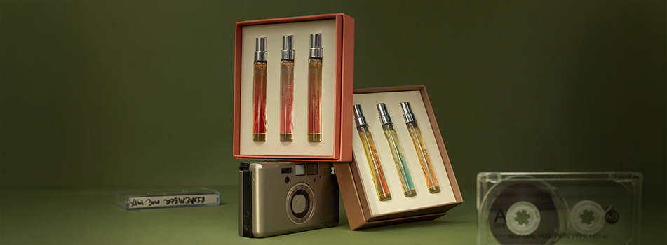 Zwei geöffnete Duftsets von Molton Brown mit jeweils vier schlanken Parfum-Miniaturen stehen auf einem olivgrünen Hintergrund. Eine Box ist auf einer silbernen Kompaktkamera aufgestellt, die andere liegt schräg daneben. Im Vorder- und Hintergrund befinden sich transparente Audiokassetten. Die Szene wirkt stilvoll, retro-inspiriert und hochwertig.