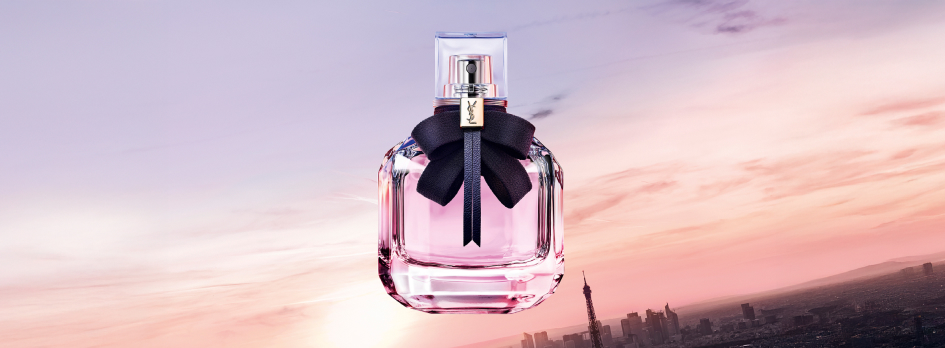 Parfumflakon von Yves Saint Laurent mit rosa Inhalt und schwarzer Schleife vor dem Schriftzug 'YSL'. Der Flakon schwebt über einer Stadtansicht mit dem Eiffelturm bei Sonnenuntergang, der Himmel ist in sanften Rosa- und Lilatönen gefärbt.