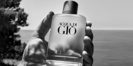 Schwarz-weiß-Fotografie einer Hand, die eine Flasche 'Acqua di Giò' Eau de Toilette von Giorgio Armani hält. Die matte Flasche mit schwarzem Verschluss trägt die Aufschrift 'Acqua di Giò – Giorgio Armani'. Im Hintergrund ist das Meer und ein Baum am Rand eines felsigen Ufers zu sehen.