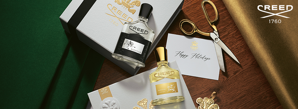 Geschenkset der Marke Creed mit zwei Parfumflakons, Aventus und Aventus for Her, auf festlicher Holzoberfläche mit Geschenkboxen, goldener Schere, Schleifenband und Karte mit der Aufschrift ‚Happy Holidays‘.