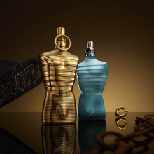 Zwei Parfumflakons in Form eines männlichen Torsos von Jean Paul Gaultier. Der linke Flakon ist goldfarben mit Ring um den Hals, der rechte ist türkisblau mit silbernem Sprühkopf. Beide stehen vor einem warm gold-braunen Hintergrund mit spiegelnder Oberfläche. Im Vordergrund liegen goldene Accessoires.