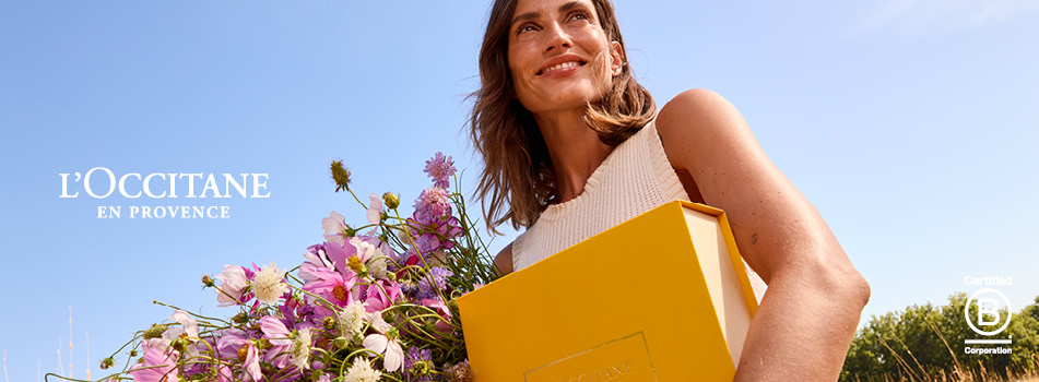 Lächelnde Frau im Sonnenlicht mit einem gelben L'OCCITANE Geschenkkarton und Blumenstrauß.