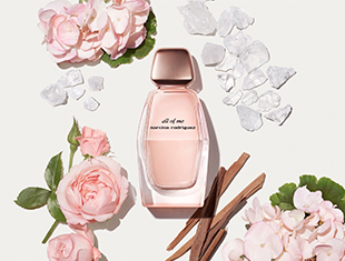 Zart roséfarbener Parfumflakon mit der Aufschrift 'all of me – narciso rodriguez' liegt auf hellem Hintergrund. Umgeben ist der Flakon von rosa Rosen, weißen Blüten, Eis- oder Kristallstücken und Holzstäbchen – symbolisch für die Duftkomponenten des Parfums.
